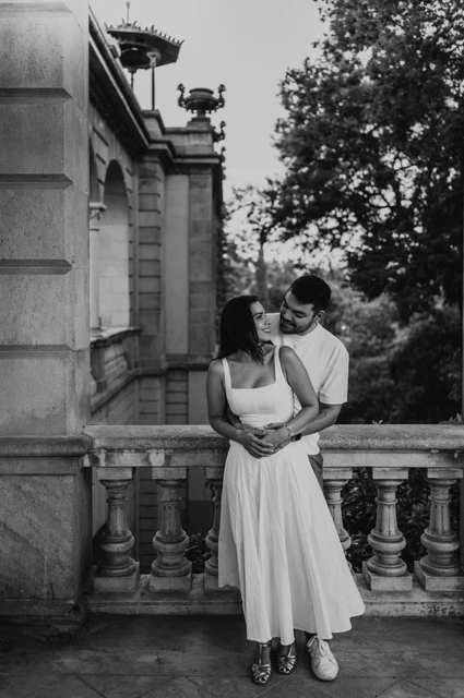 A quiet embrace under the arch in black and white | Barcelona engagement photoshoot Black-and-white photograph: the couple leaning close together under a stone arch with greenery beyond the railing during their engagement photoshoot in Barcelona.