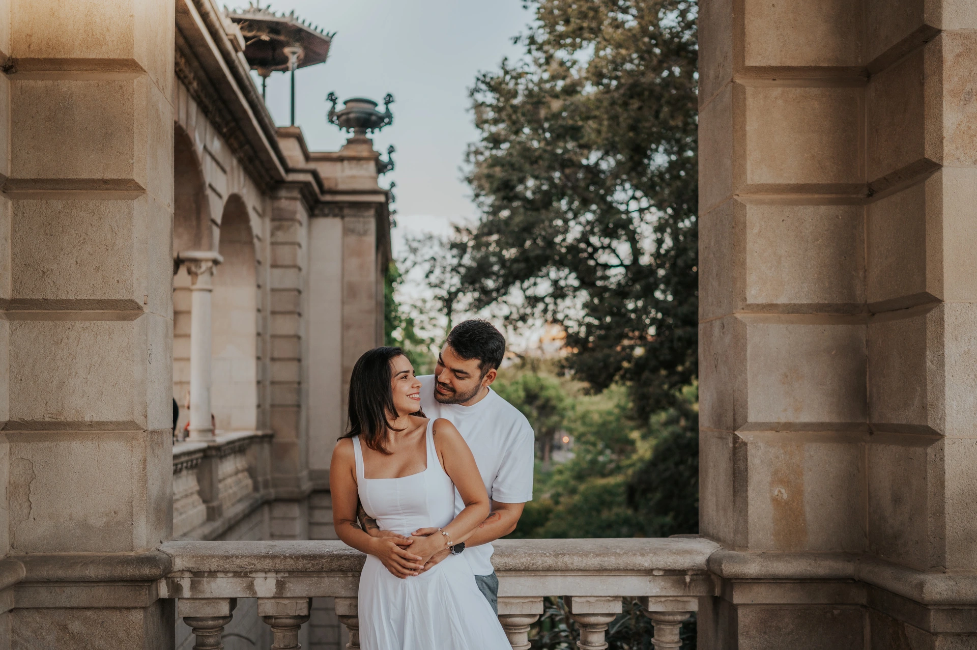 The couple leaning close together under a stone arch with greenery beyond the railing during their engagement photoshoot in Barcelona.