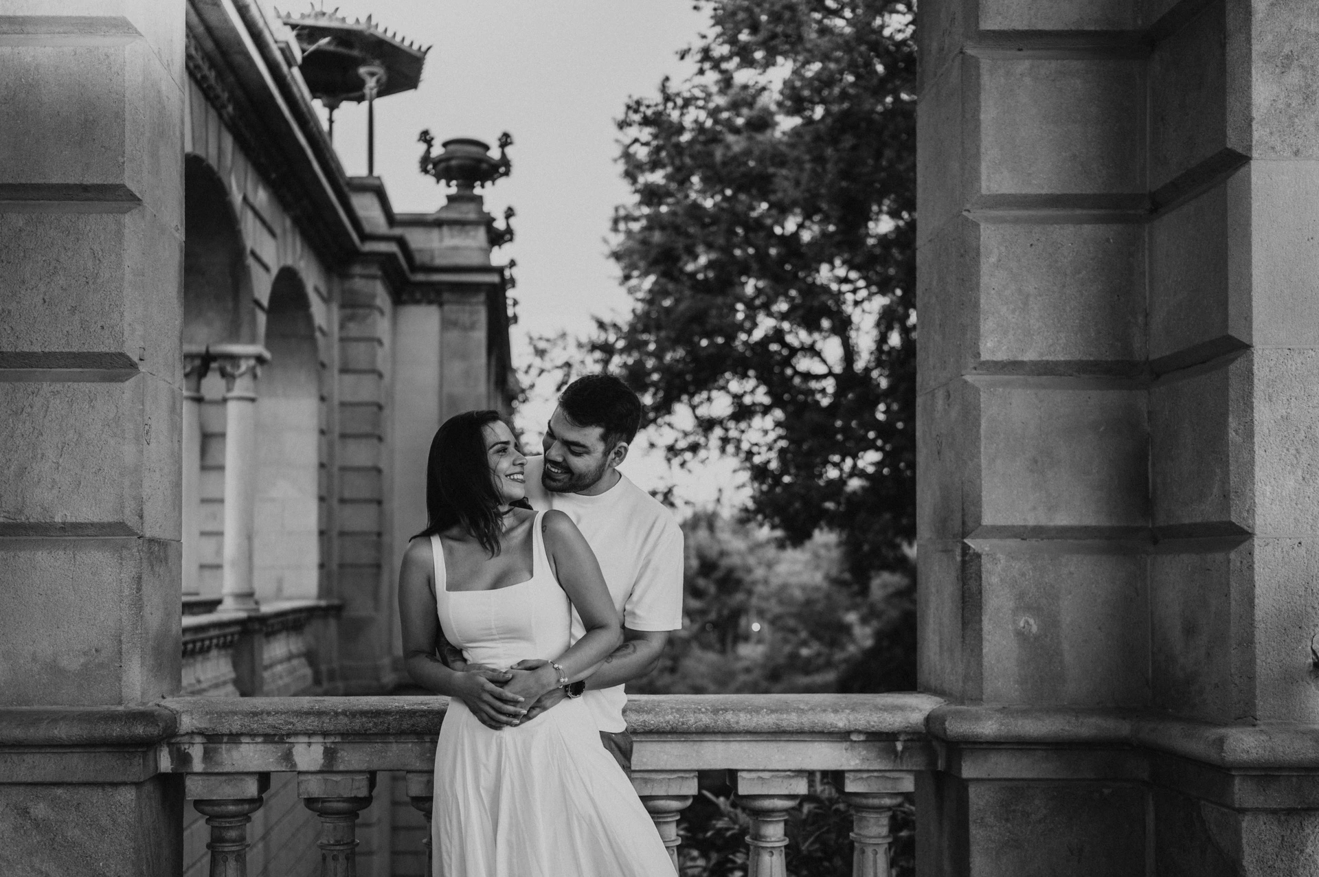 Black-and-white photograph: the couple leaning close together under a stone arch with greenery beyond the railing during their engagement photoshoot in Barcelona.