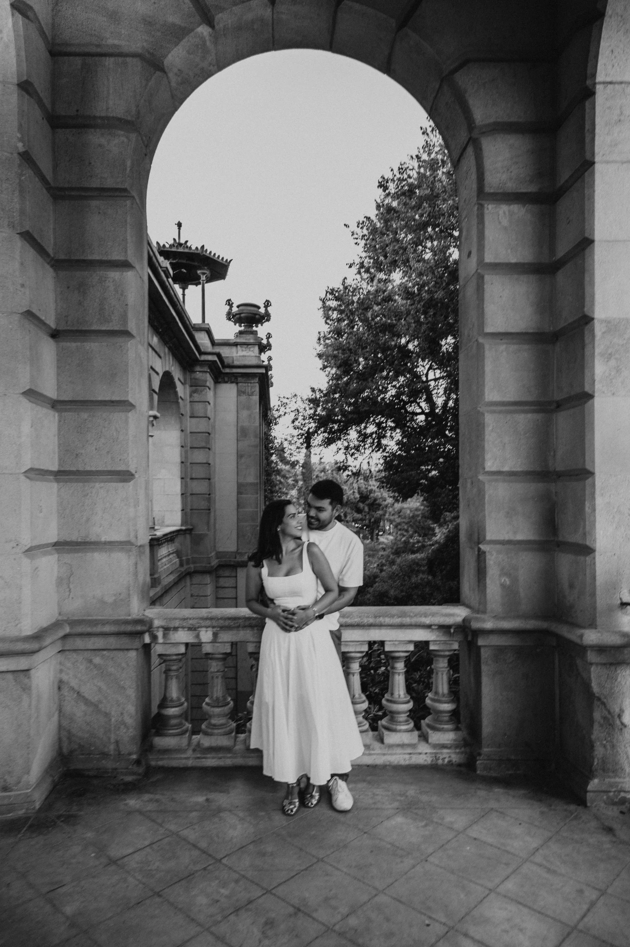 Black-and-white photograph: a full-length portrait of the couple holding each other in a tall stone archway during their engagement photoshoot in Barcelona.