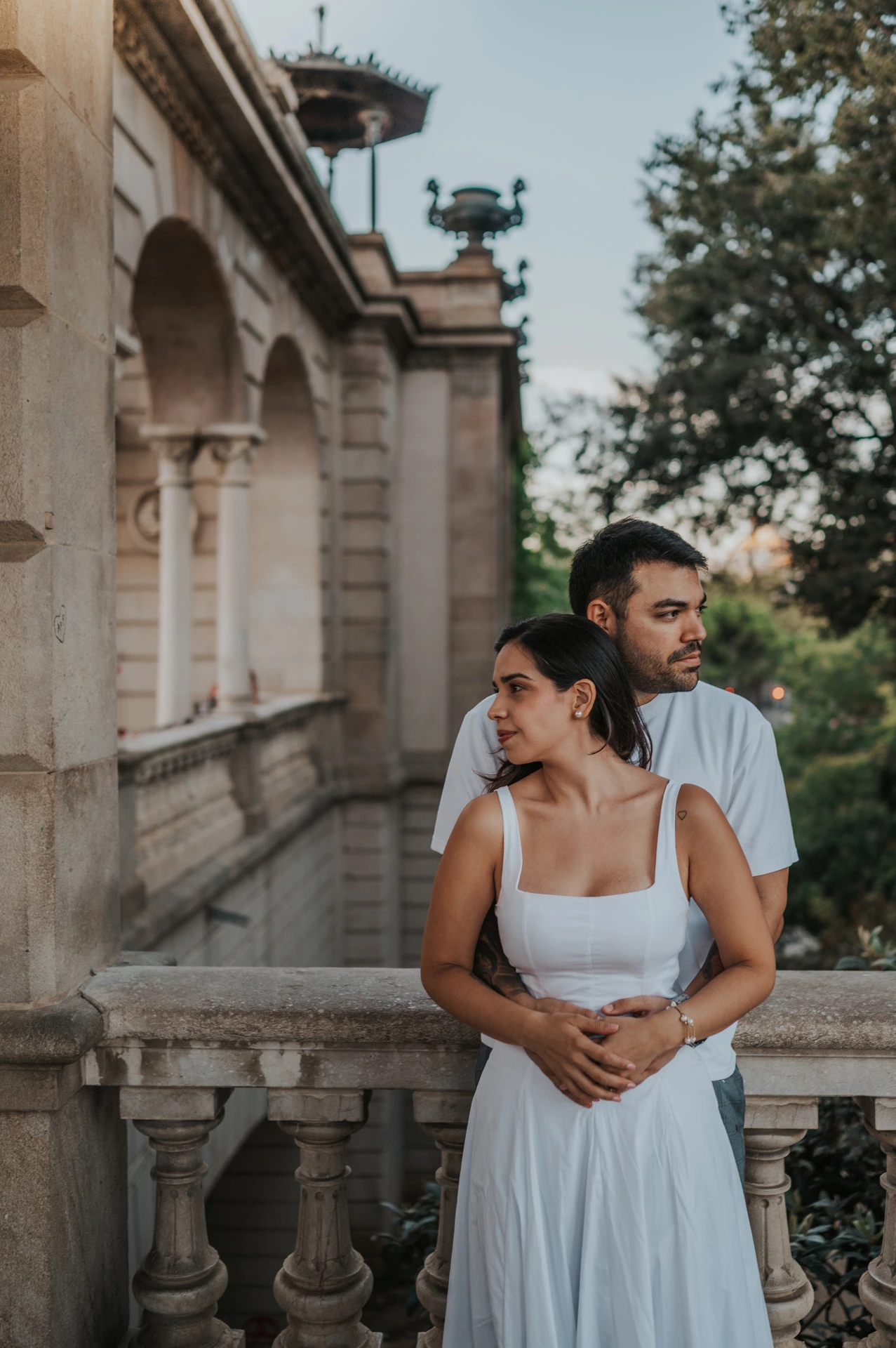The couple embracing beside a stone pillar and looking out in different directions during their engagement photoshoot in Barcelona.