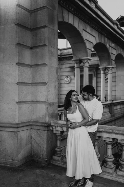 Together along the arched walkway in black and white | Barcelona engagement photoshoot Black-and-white photograph: the couple standing close together beside the stone arches and balustrade during their engagement photoshoot in Barcelona.