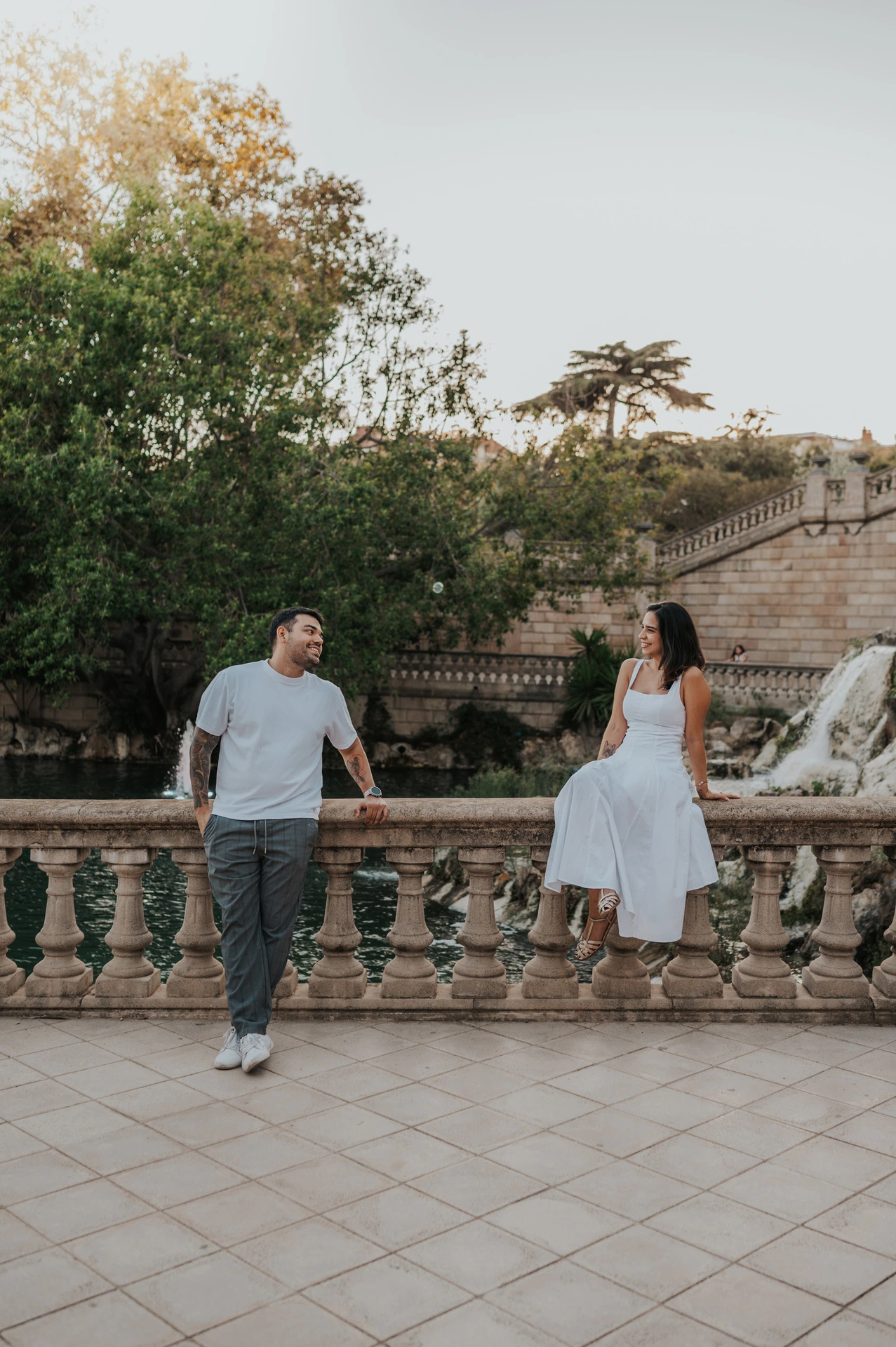 The couple holding hands across a stone balustrade, with the woman sitting on its edge during their engagement photoshoot in Barcelona.
