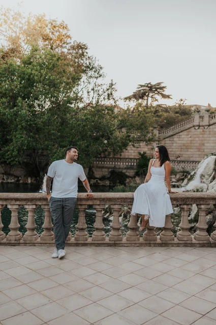 A playful portrait along the stone railing | Barcelona engagement photoshoot The couple holding hands across a stone balustrade, with the woman sitting on its edge during their engagement photoshoot in Barcelona.