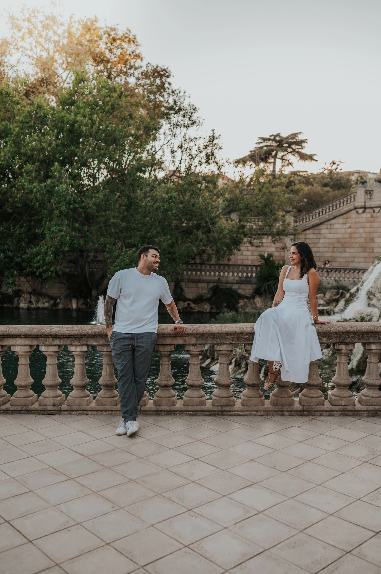 The couple holding hands across a stone balustrade, with the woman sitting on its edge during their engagement photoshoot in Barcelona.