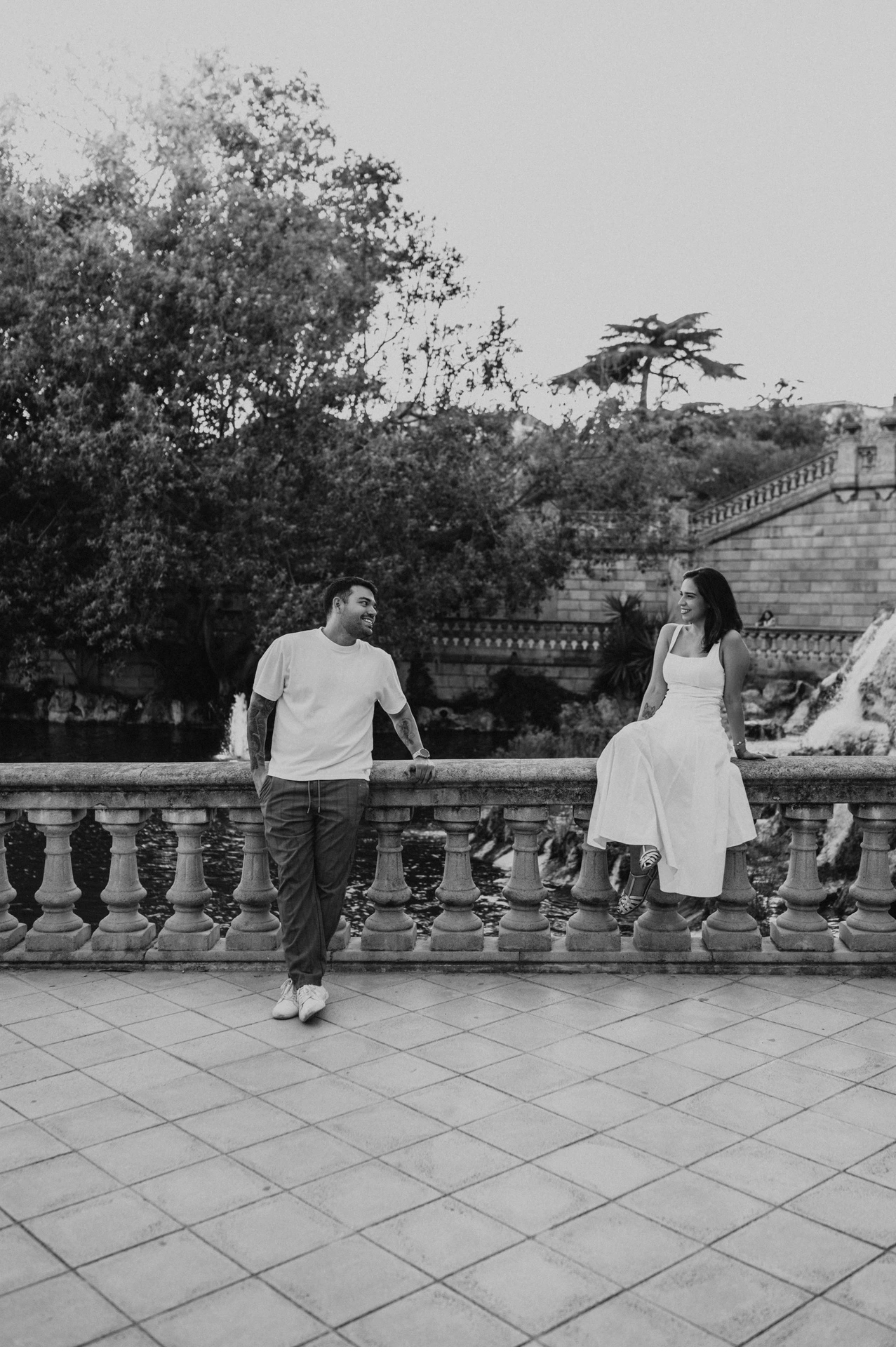 Black-and-white photograph: the couple holding hands across a stone balustrade, with the woman sitting on its edge during their engagement photoshoot in Barcelona.