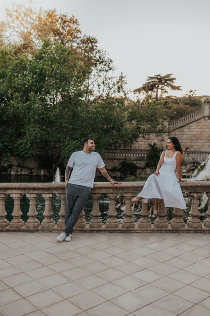 A playful portrait along the stone railing | Barcelona engagement photoshoot The couple holding hands across a stone balustrade, with the woman sitting on its edge during their engagement photoshoot in Barcelona.