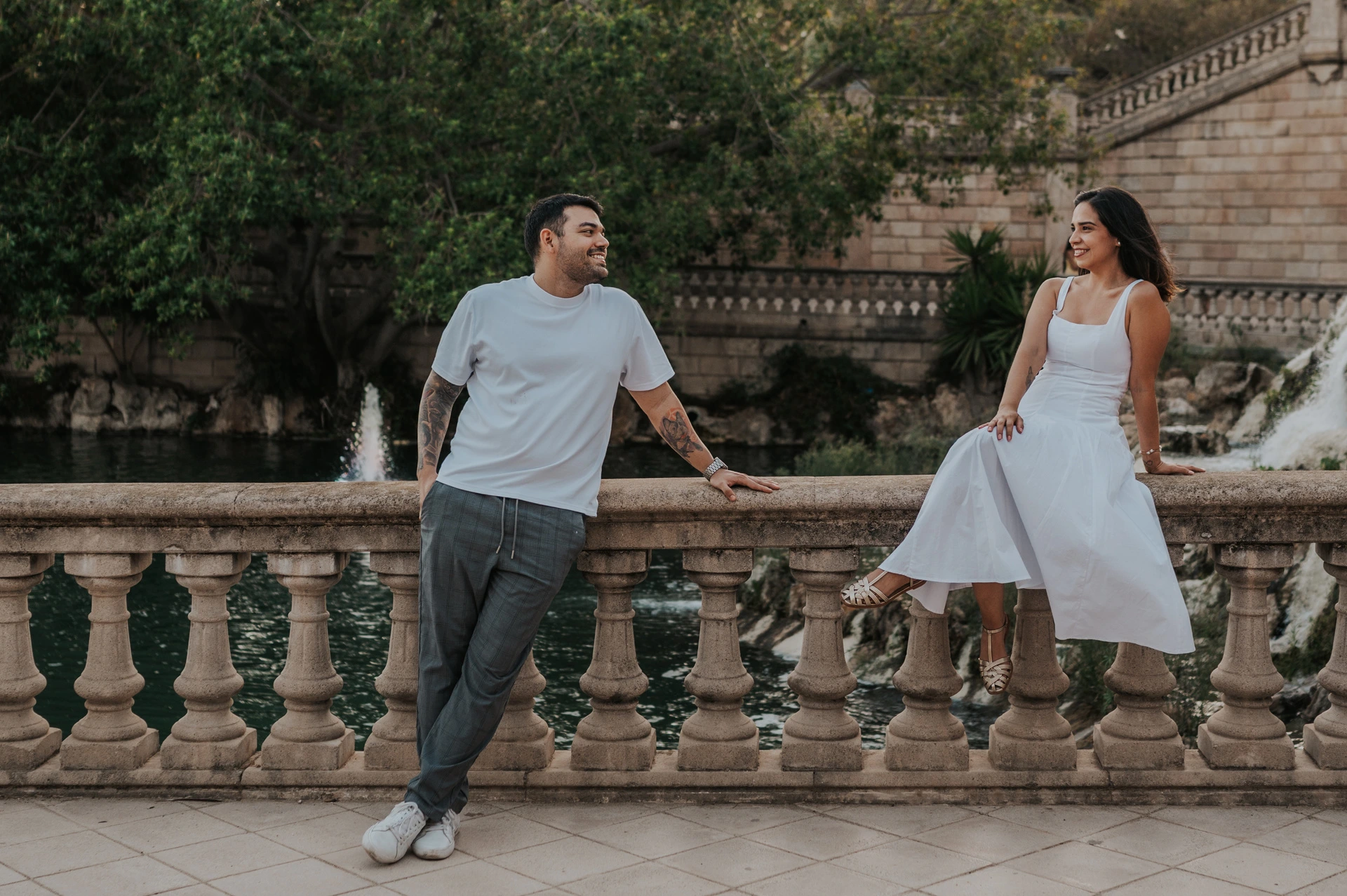 A playful portrait along the stone railing | Barcelona engagement photoshoot The couple holding hands across a stone balustrade, with the woman sitting on its edge during their engagement photoshoot in Barcelona.