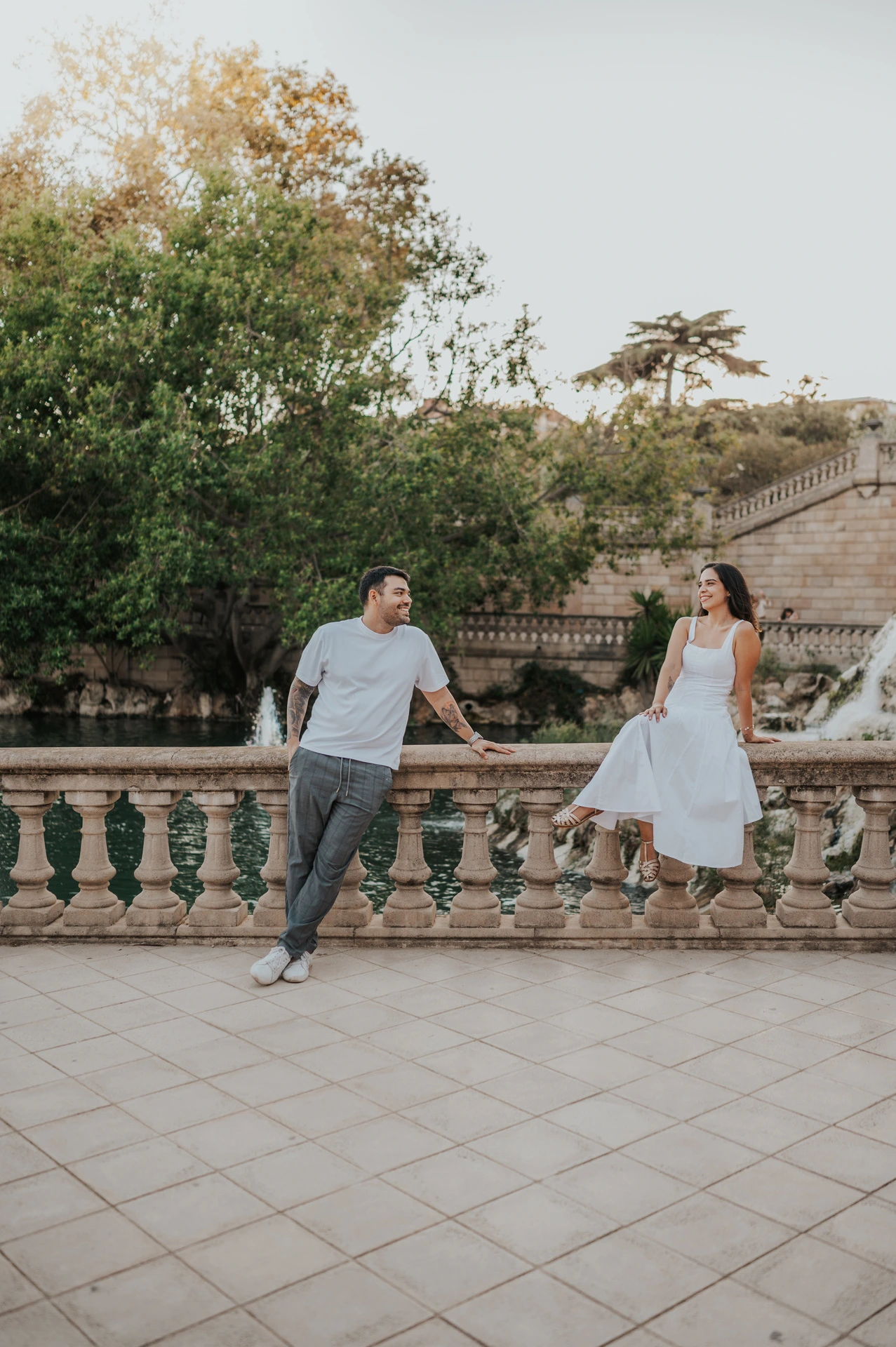 The couple holding hands across a stone balustrade, with the woman sitting on its edge during their engagement photoshoot in Barcelona.
