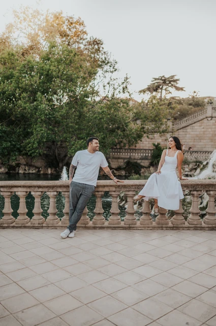 A playful portrait along the stone railing | Barcelona engagement photoshoot The couple holding hands across a stone balustrade, with the woman sitting on its edge during their engagement photoshoot in Barcelona.