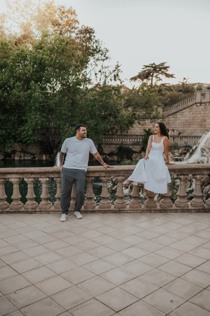 A playful portrait along the stone railing | Barcelona engagement photoshoot The couple holding hands across a stone balustrade, with the woman sitting on its edge during their engagement photoshoot in Barcelona.