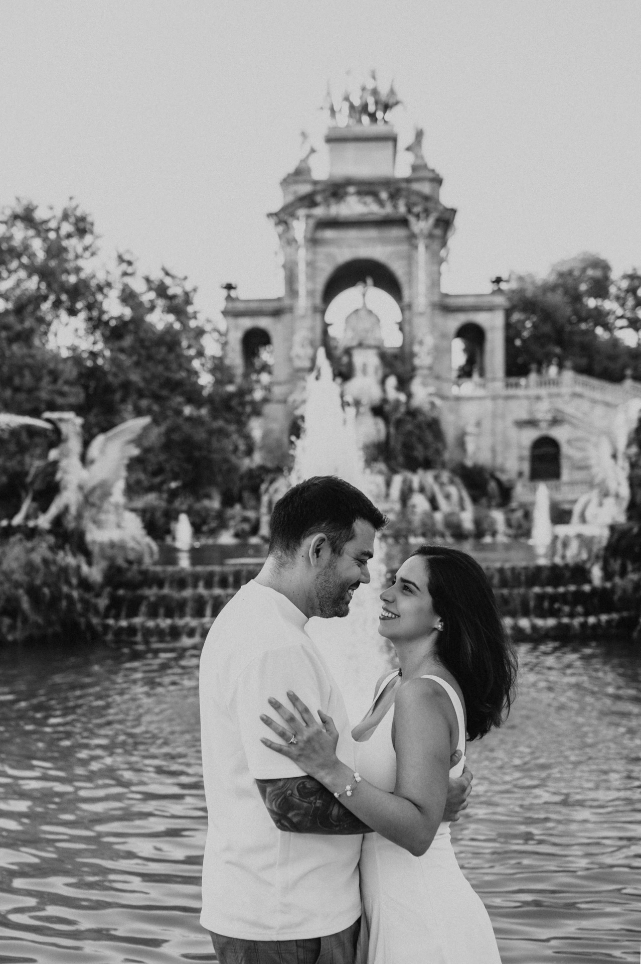 Black-and-white photograph: the couple facing each other in front of the fountain and its golden-topped arches during their engagement photoshoot in Barcelona.