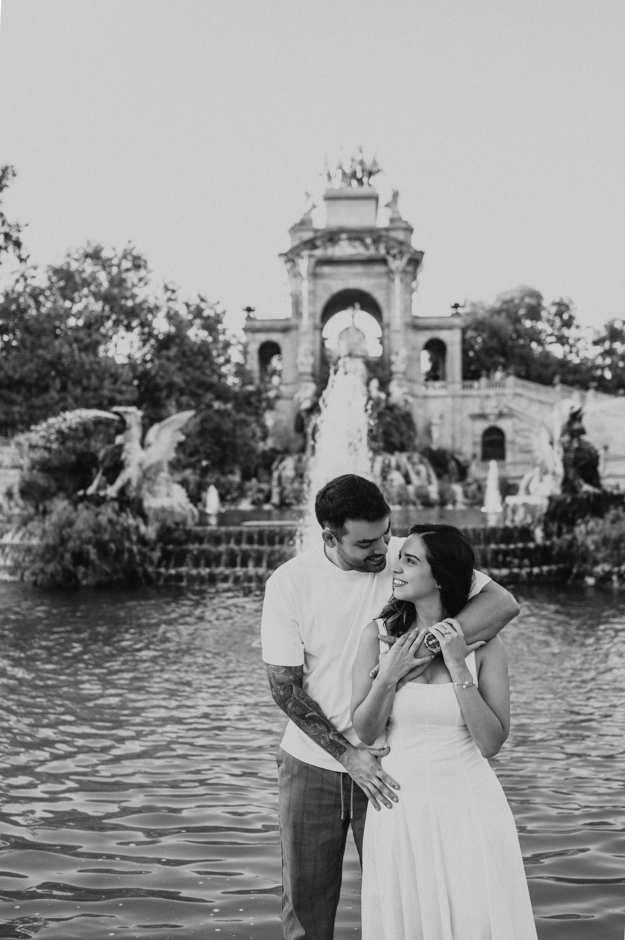 Black-and-white photograph: the man with his arm around his partner as they stand together beside the fountain pool during their engagement photoshoot in Barcelona.