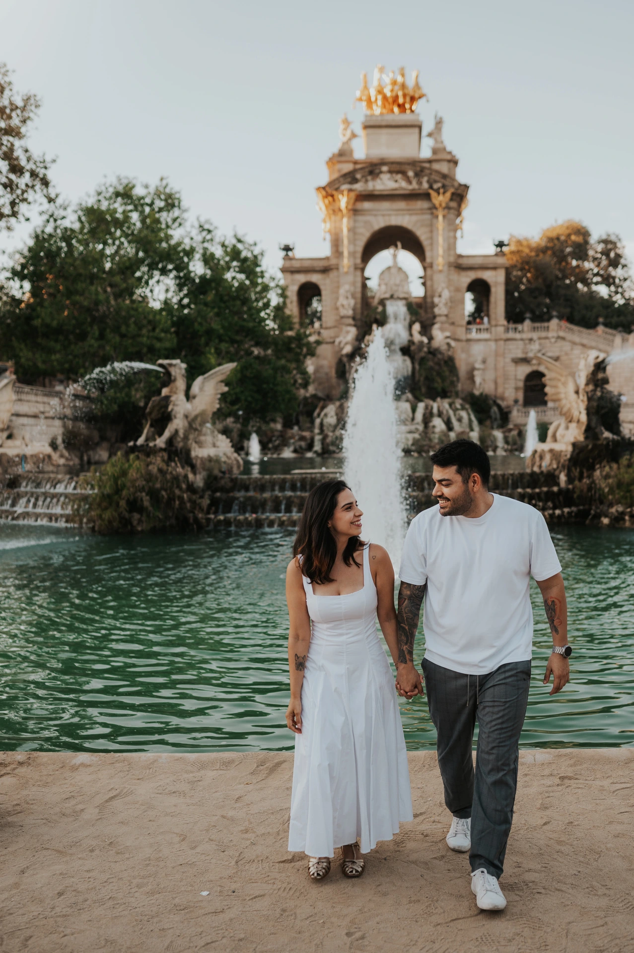 The couple walking hand in hand beside the green fountain pool, with the ornate monument behind them during their engagement photoshoot in Barcelona.