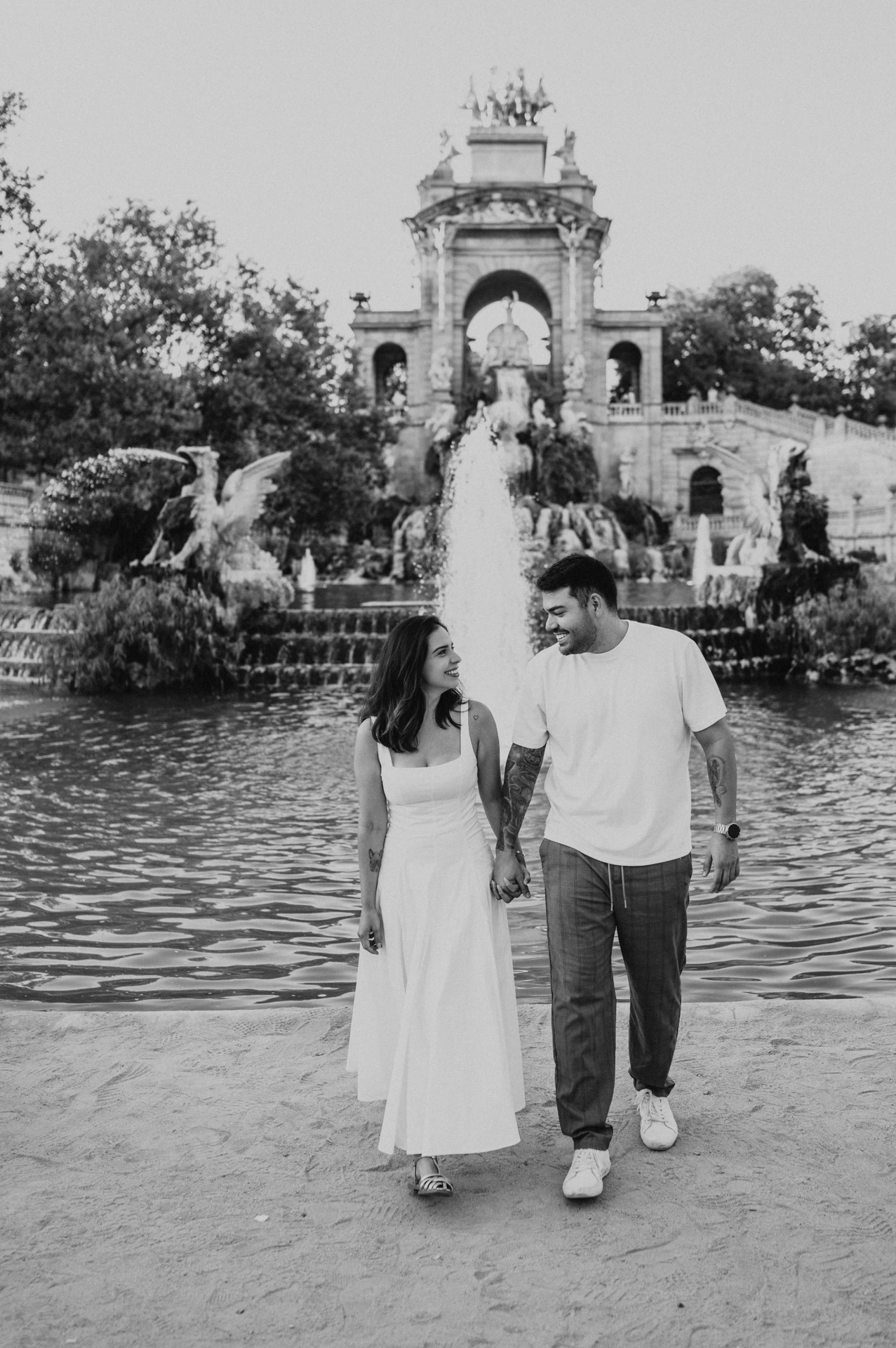 Black-and-white photograph: the couple walking hand in hand beside the green fountain pool, with the ornate monument behind them during their engagement photoshoot in Barcelona.