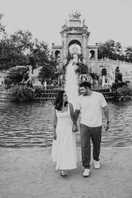 A walk beside the fountain pool in black and white | Barcelona engagement photoshoot Black-and-white photograph: the couple walking hand in hand beside the green fountain pool, with the ornate monument behind them during their engagement photoshoot in Barcelona.