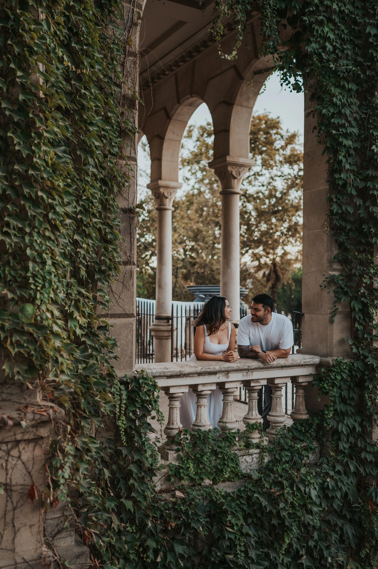 The couple leaning towards each other behind a stone balustrade framed by ivy-covered arches during their engagement photoshoot in Barcelona.