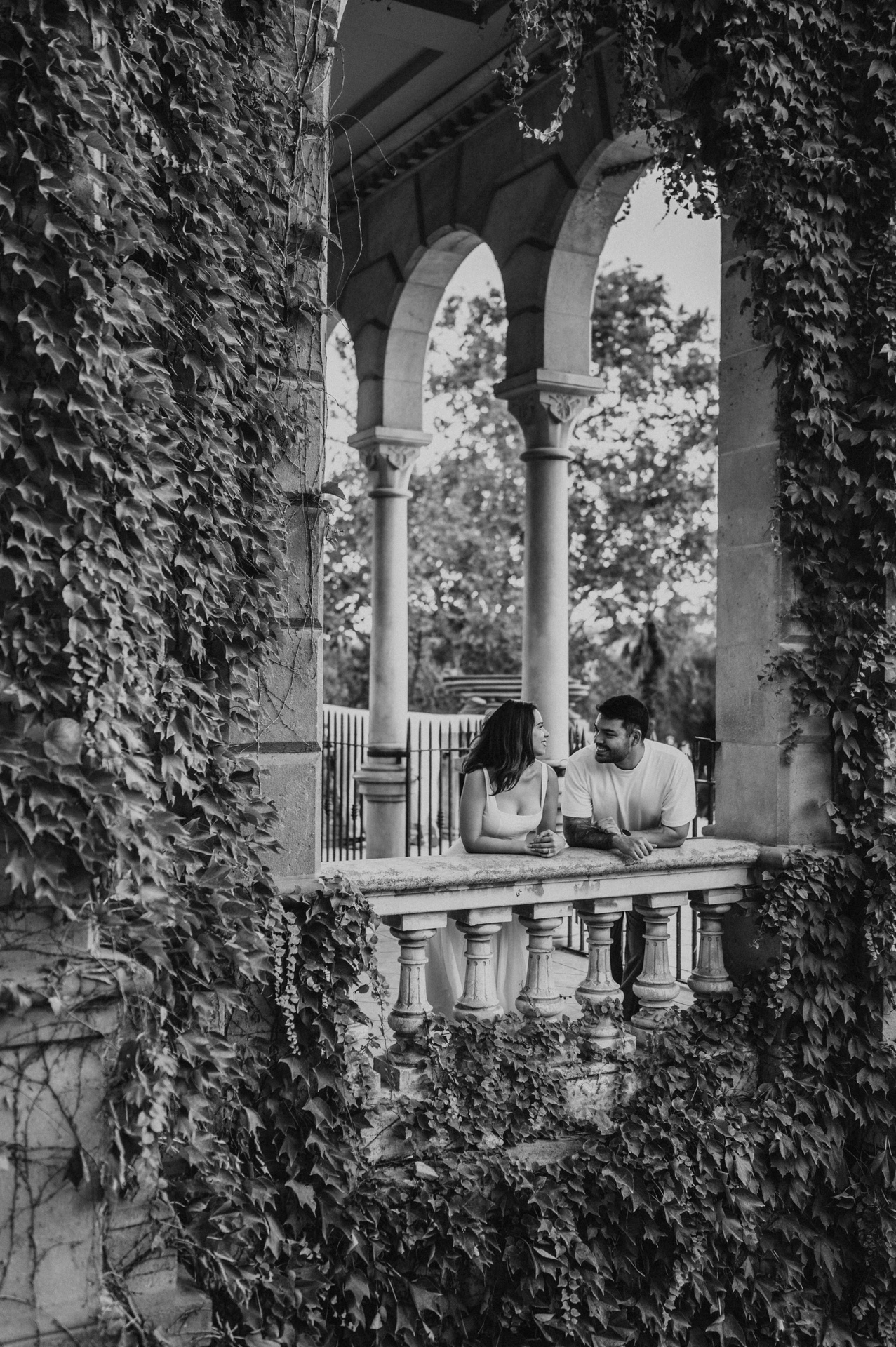 Black-and-white photograph: the couple leaning towards each other behind a stone balustrade framed by ivy-covered arches during their engagement photoshoot in Barcelona.