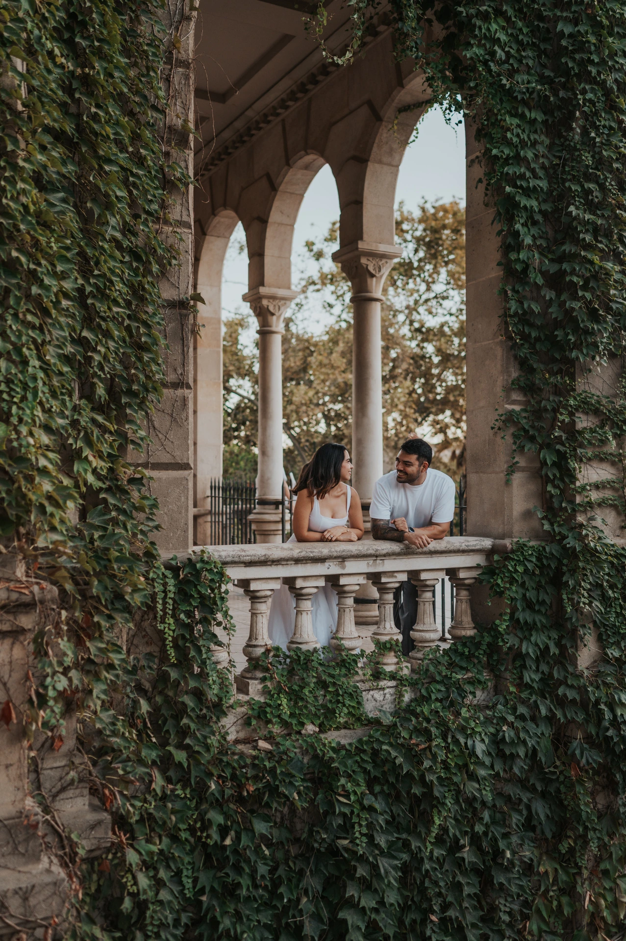 The couple leaning towards each other behind a stone balustrade framed by ivy-covered arches during their engagement photoshoot in Barcelona.