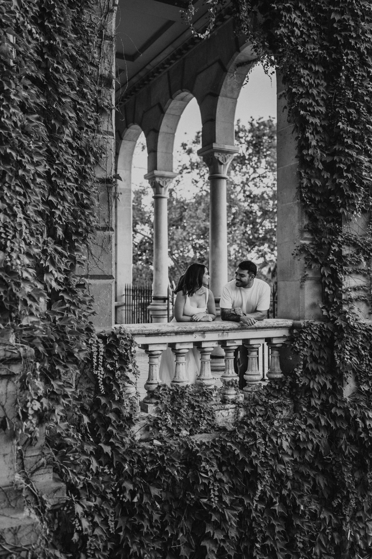 Black-and-white photograph: the couple leaning towards each other behind a stone balustrade framed by ivy-covered arches during their engagement photoshoot in Barcelona.