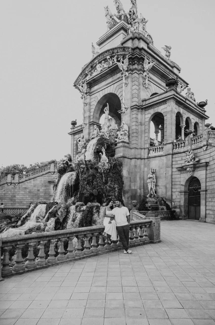 The fountain monument in a wide portrait in black and white | Barcelona engagement photoshoot Black-and-white photograph: a wide view of the couple beside the fountain’s stone railing and monumental arches during their engagement photoshoot in Barcelona.