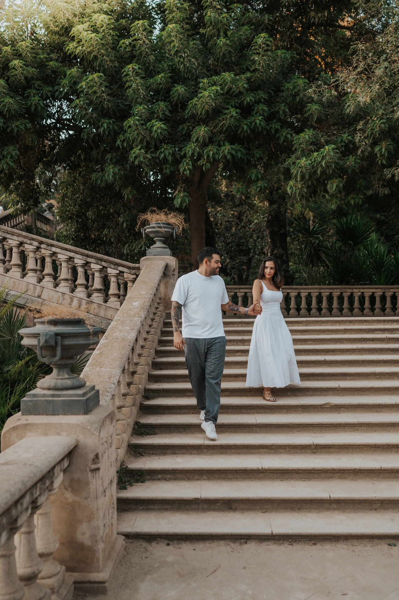 The couple walking down the broad stone staircase, holding hands beneath leafy trees during their engagement photoshoot in Barcelona.