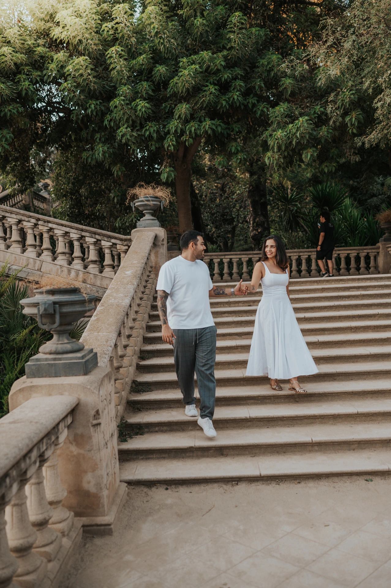 The couple walking down the broad stone staircase, holding hands beneath leafy trees during their engagement photoshoot in Barcelona.
