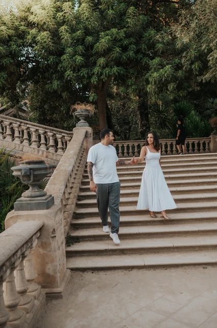The couple walking down the broad stone staircase, holding hands beneath leafy trees during their engagement photoshoot in Barcelona.