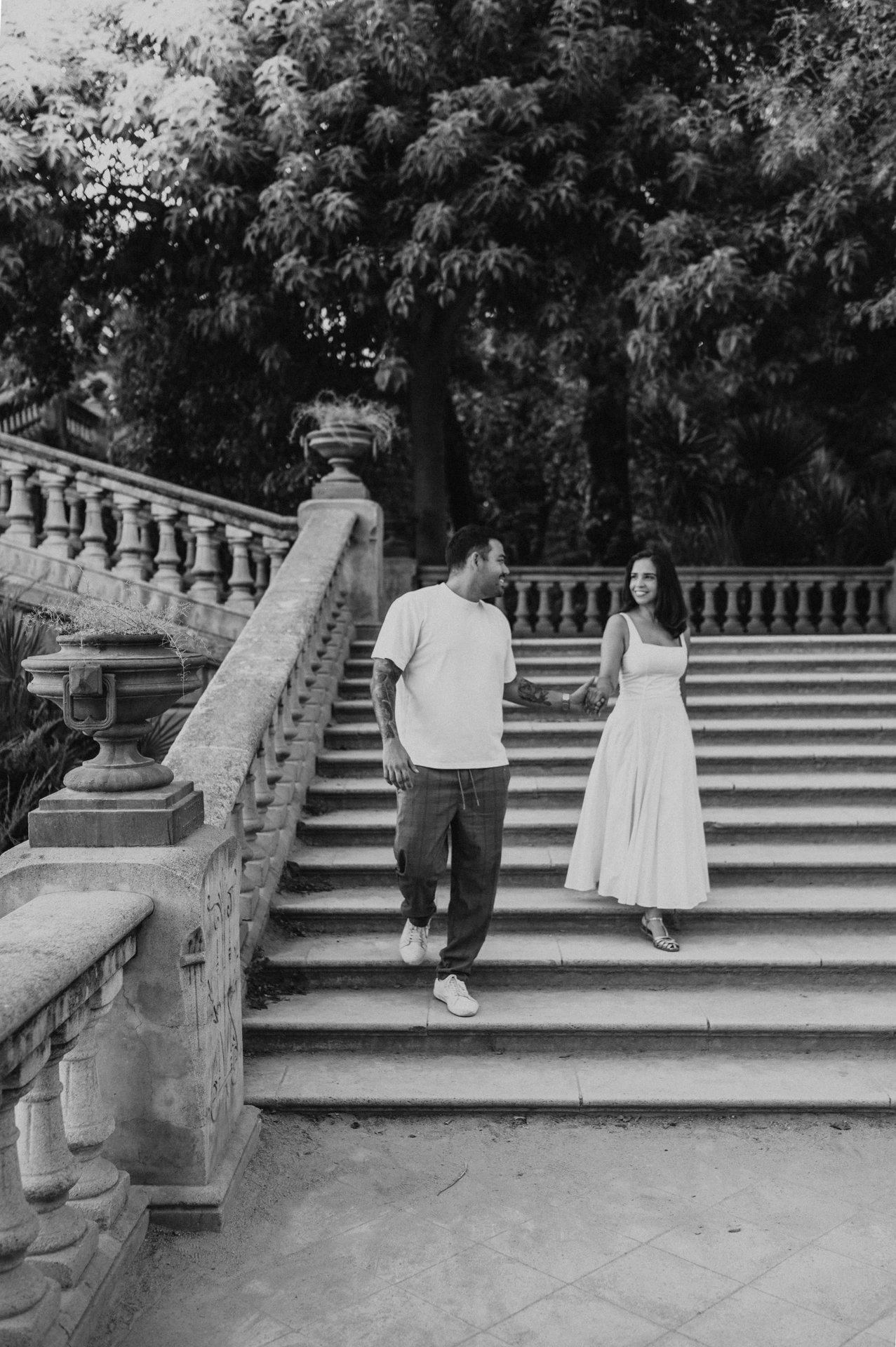 Black-and-white photograph: the couple walking down the broad stone staircase, holding hands beneath leafy trees during their engagement photoshoot in Barcelona.
