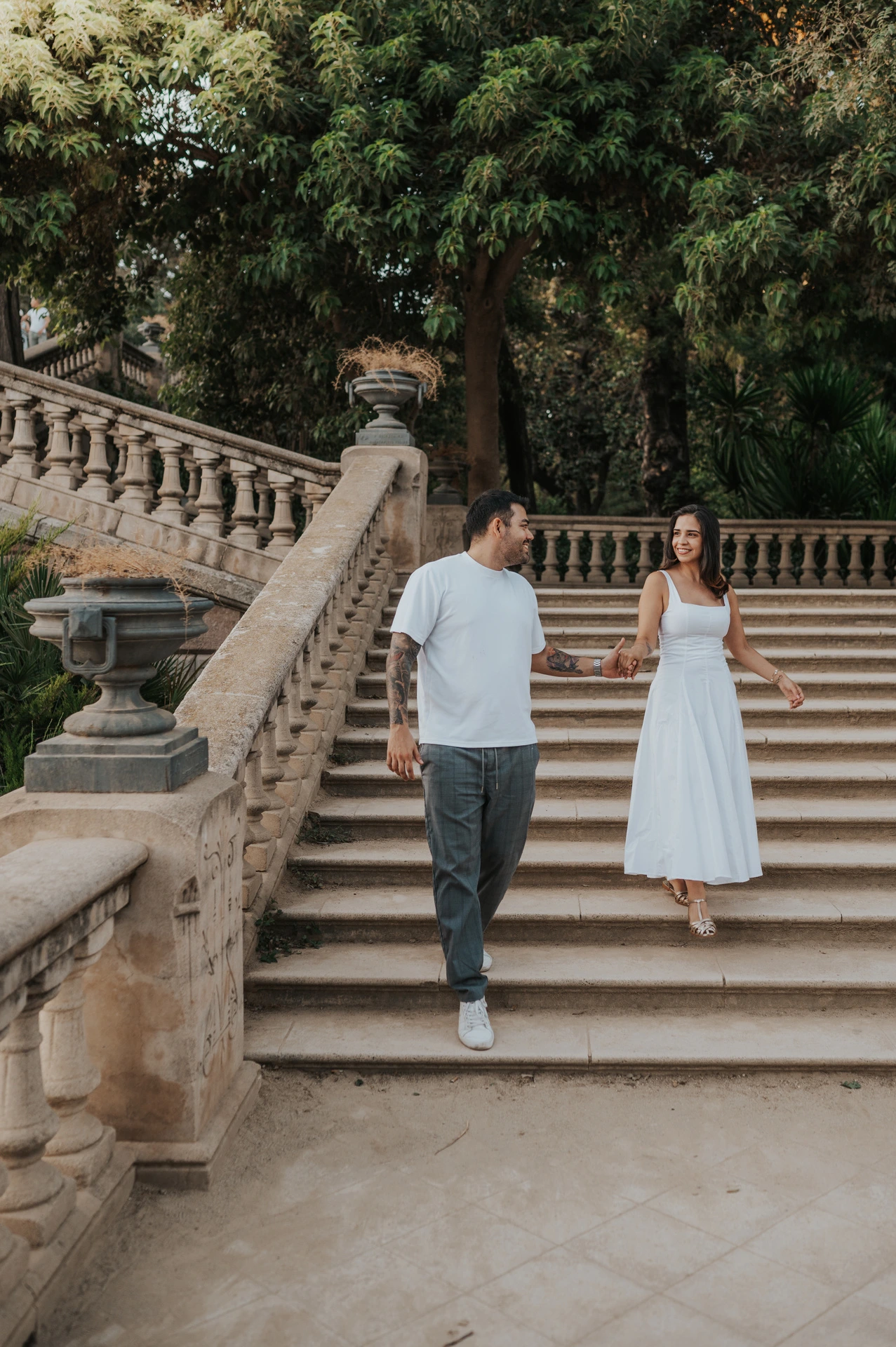 The couple walking down the broad stone staircase, holding hands beneath leafy trees during their engagement photoshoot in Barcelona.