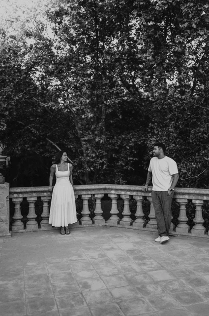 A terrace portrait beneath leafy trees in black and white | Barcelona engagement photoshoot Black-and-white photograph: the couple standing apart along a stone terrace railing, looking towards each other beneath the trees during their engagement photoshoot in Barcelona.