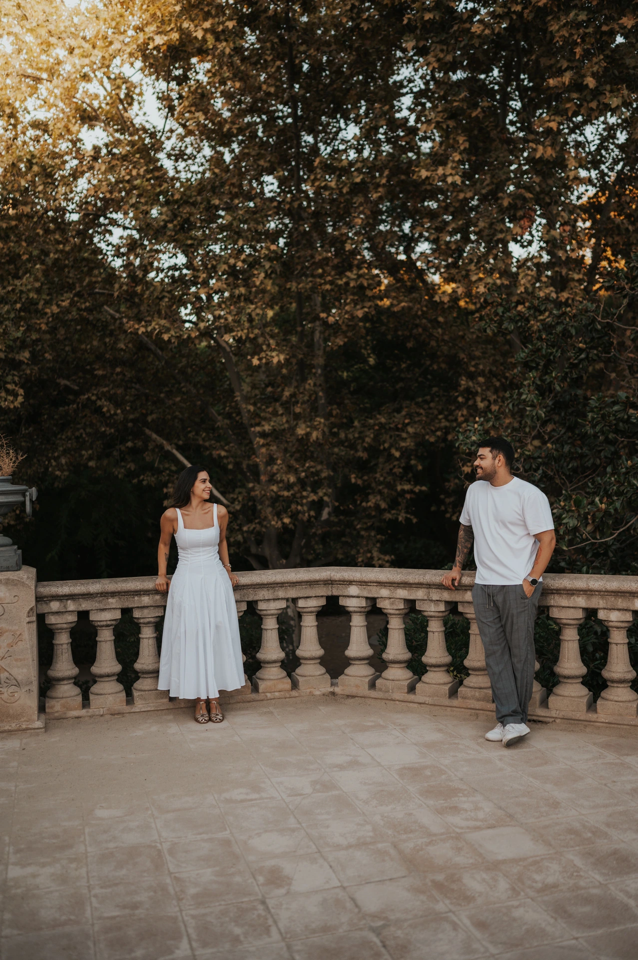 The couple standing apart along a stone terrace railing, looking towards each other beneath the trees during their engagement photoshoot in Barcelona.