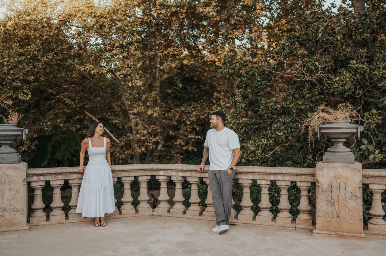 A terrace portrait beneath leafy trees | Barcelona engagement photoshoot The couple standing apart along a stone terrace railing, looking towards each other beneath the trees during their engagement photoshoot in Barcelona.