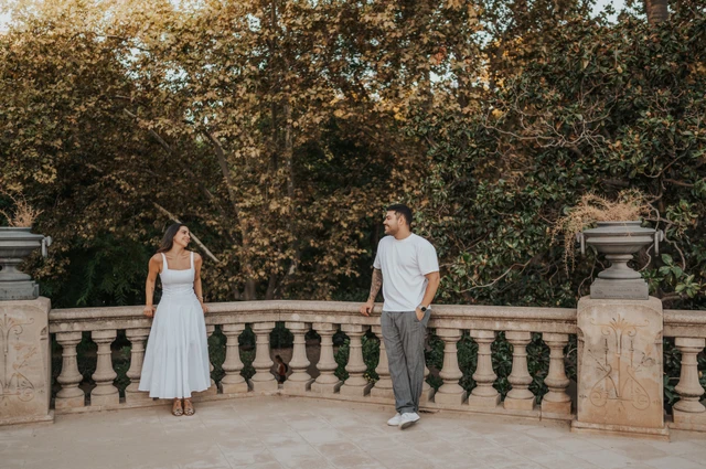The couple standing apart along a stone terrace railing, looking towards each other beneath the trees during their engagement photoshoot in Barcelona.