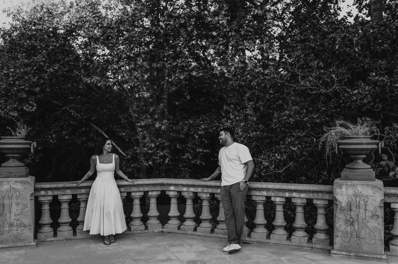 A terrace portrait beneath leafy trees in black and white | Barcelona engagement photoshoot Black-and-white photograph: the couple standing apart along a stone terrace railing, looking towards each other beneath the trees during their engagement photoshoot in Barcelona.