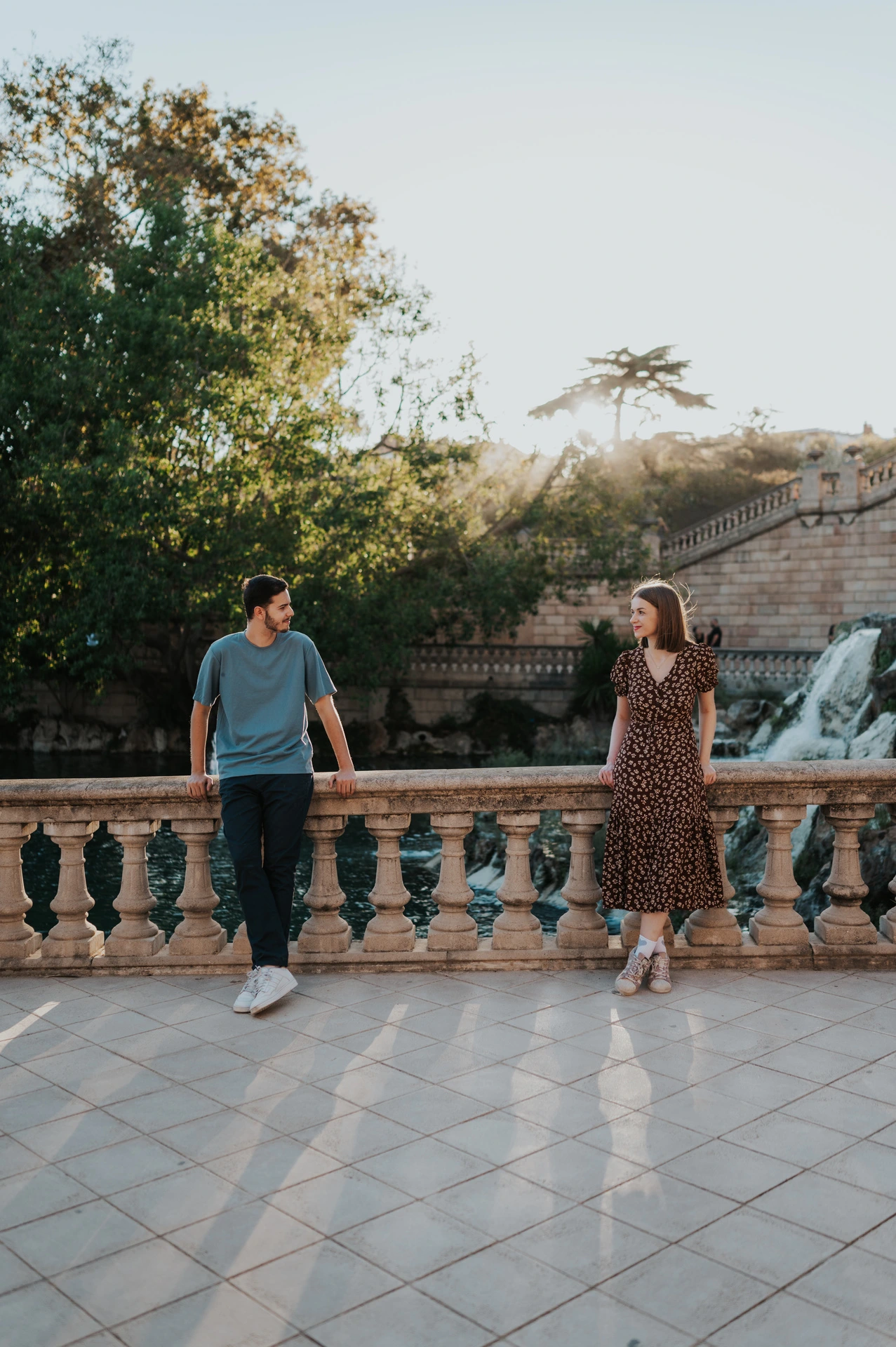 The couple standing apart along a stone balustrade, looking towards each other with trees and water behind them during their couple photoshoot in Barcelona.
