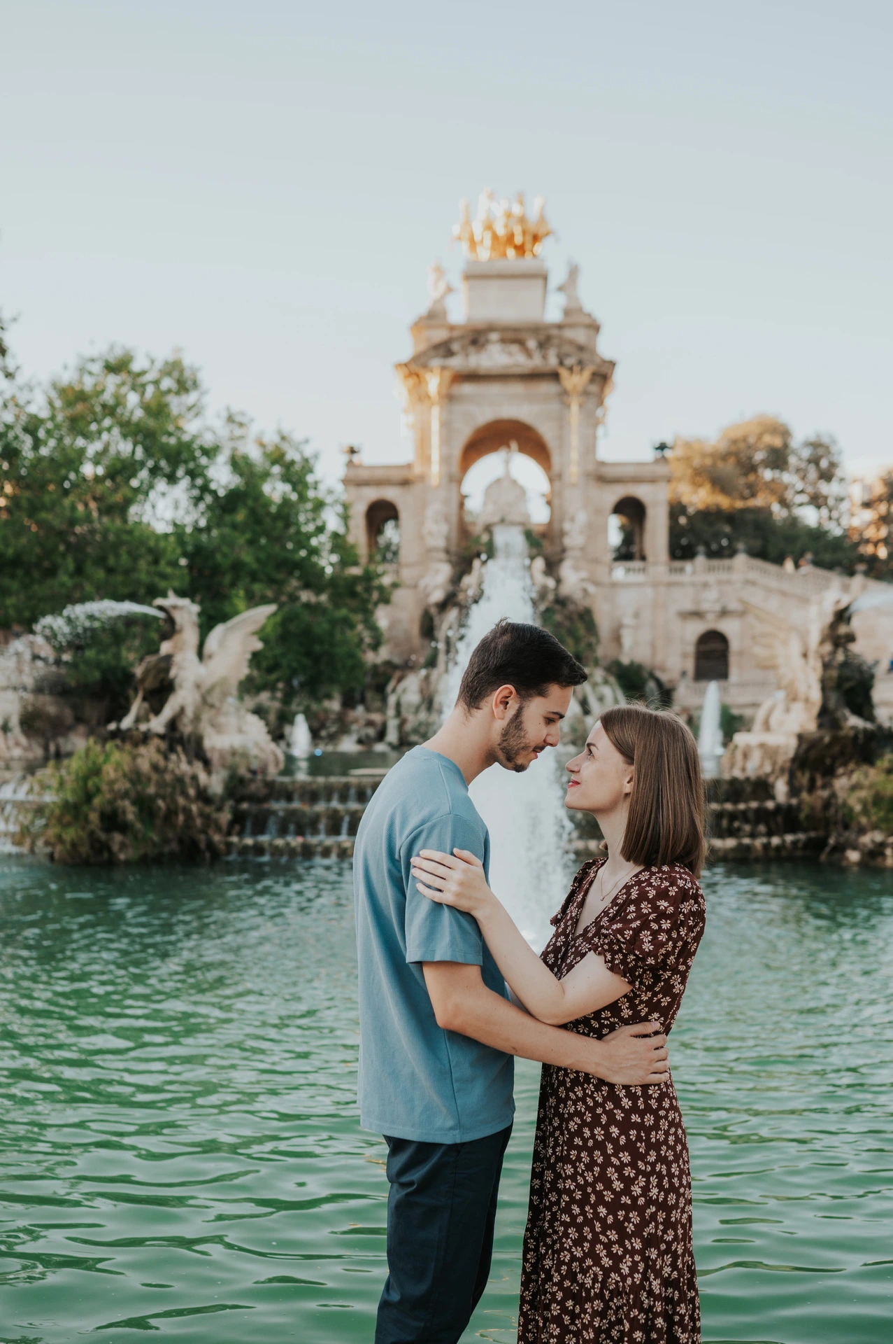 The couple facing each other beside the green fountain pool, with its golden-topped arches behind them during their couple photoshoot in Barcelona.
