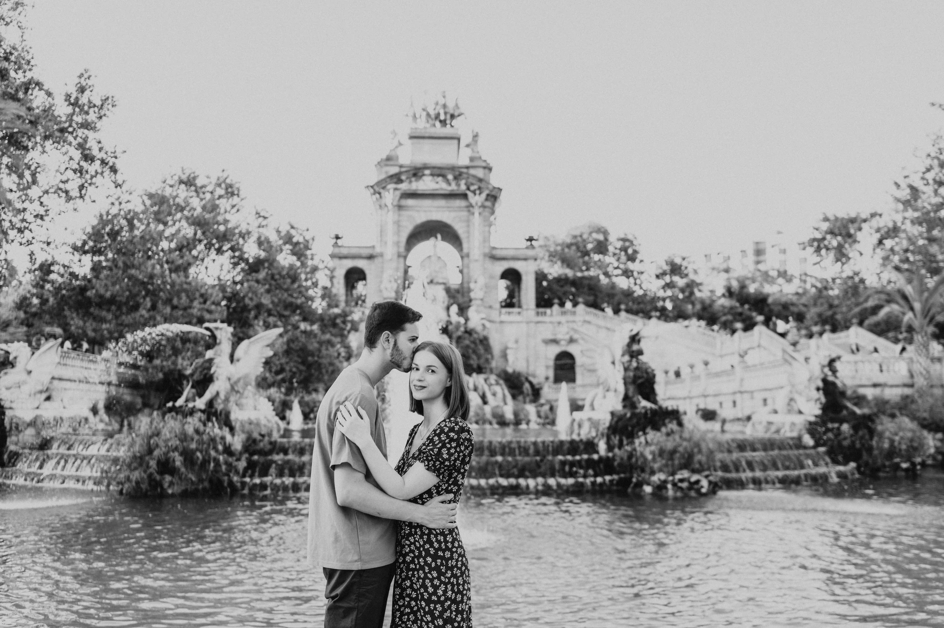 Black-and-white photograph: the couple holding each other in front of the fountain pool and ornate stone monument during their couple photoshoot in Barcelona.