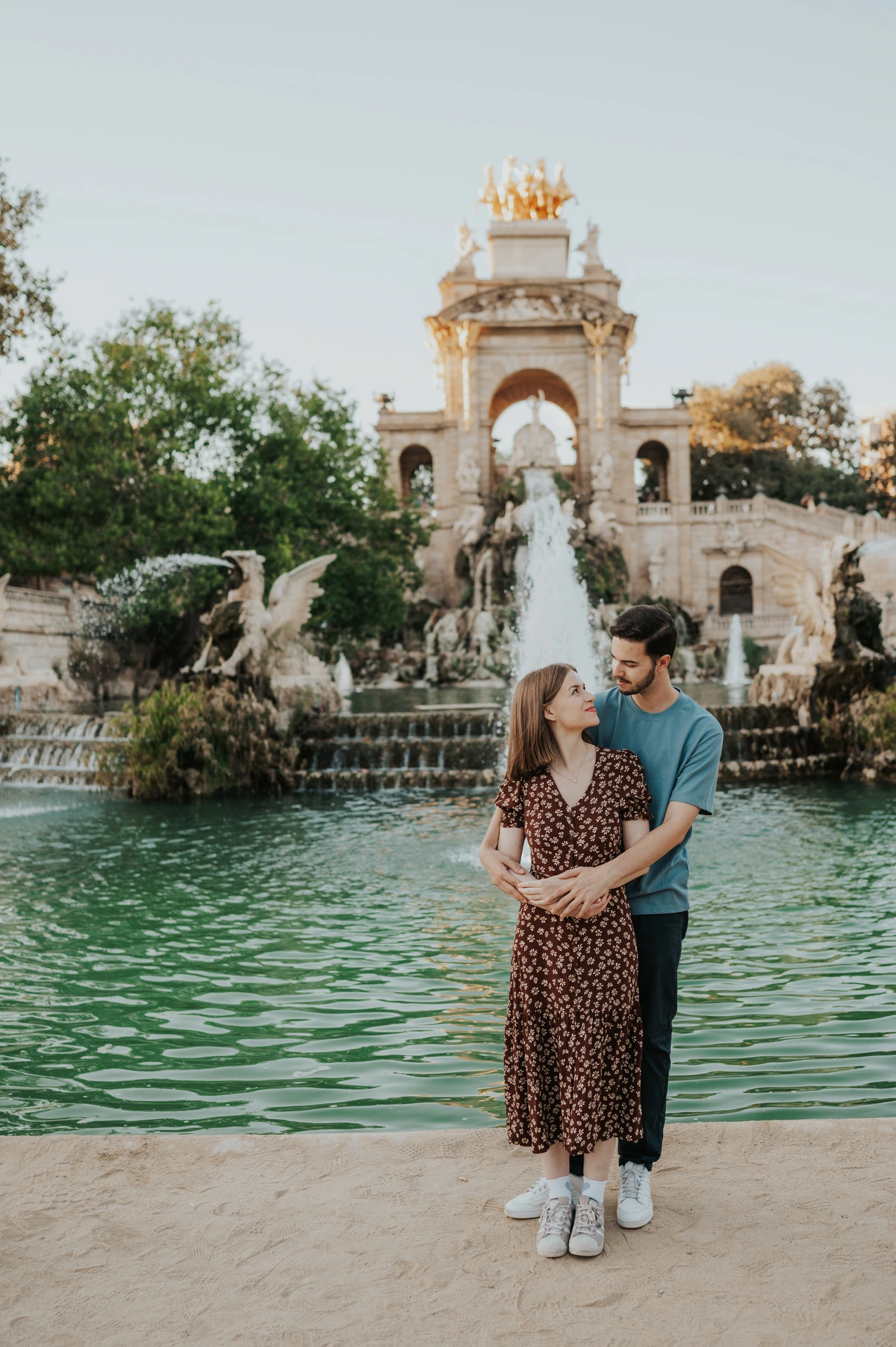 The man standing behind his partner and holding her close in front of the park fountain during their couple photoshoot in Barcelona.