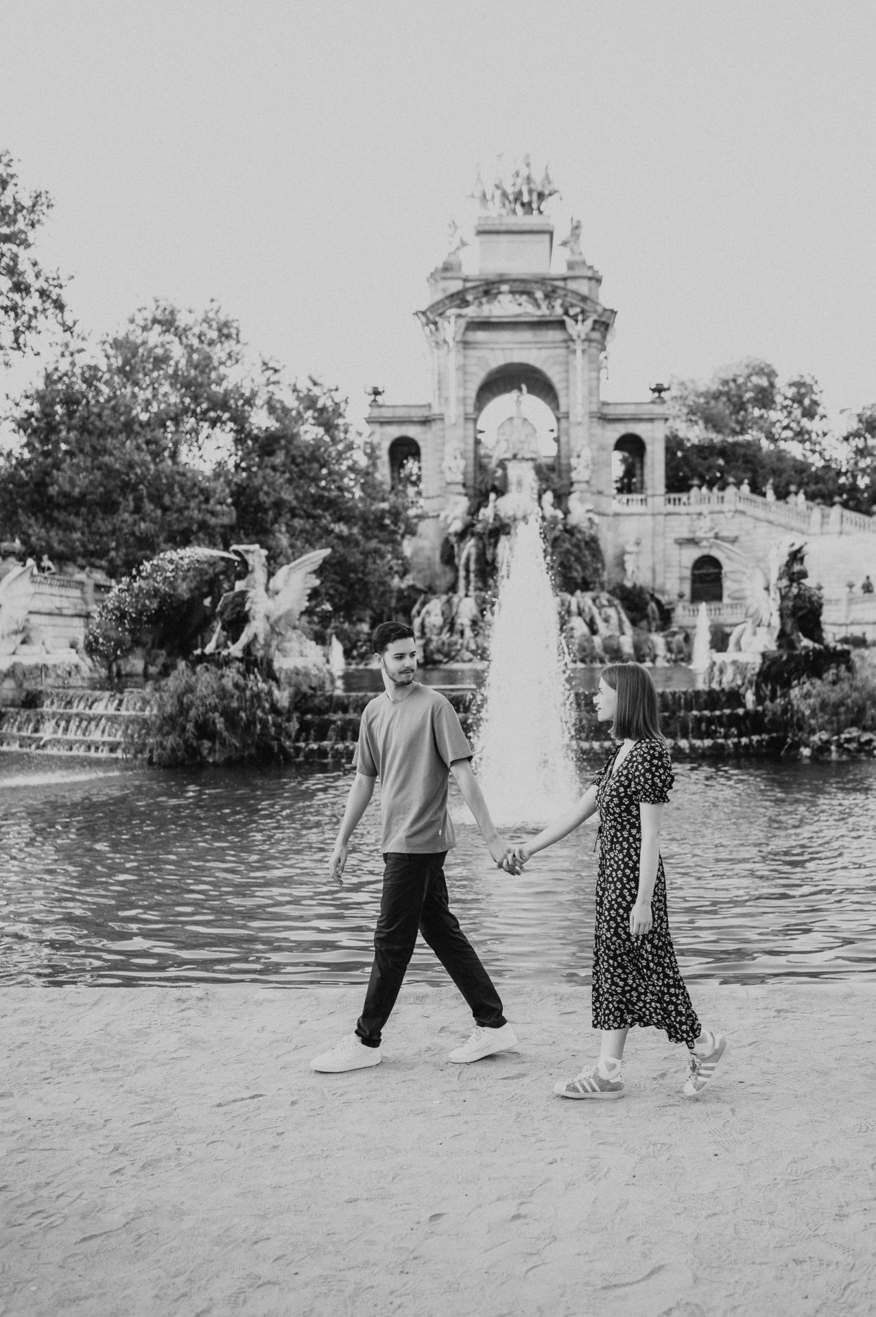 Black-and-white photograph: the couple holding hands beside the green pool, with the fountain monument in the background during their couple photoshoot in Barcelona.