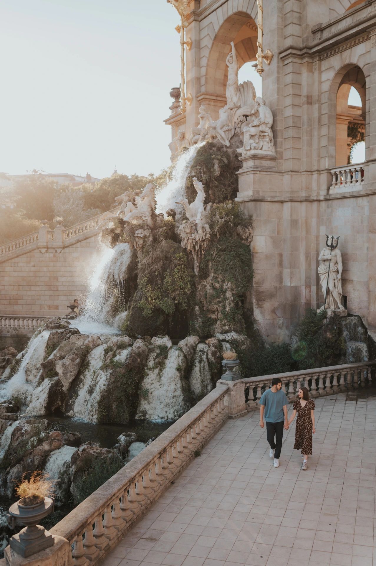 The couple walking hand in hand beside the fountain, with its ornate stone arches rising above them during their couple photoshoot in Barcelona.