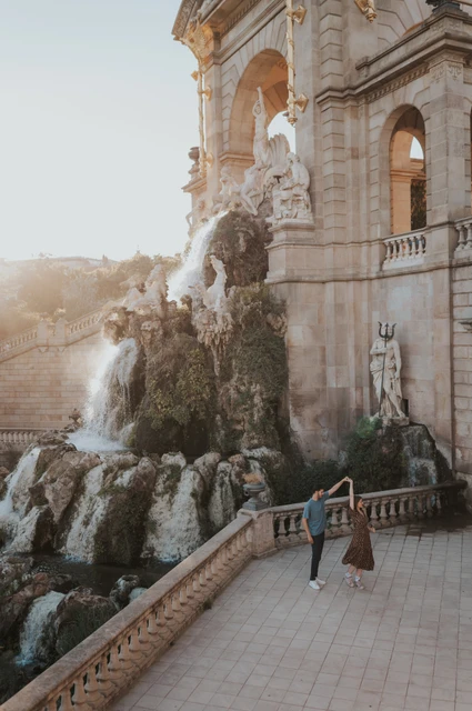 The couple walking hand in hand beside the fountain, with its ornate stone arches rising above them during their couple photoshoot in Barcelona.
