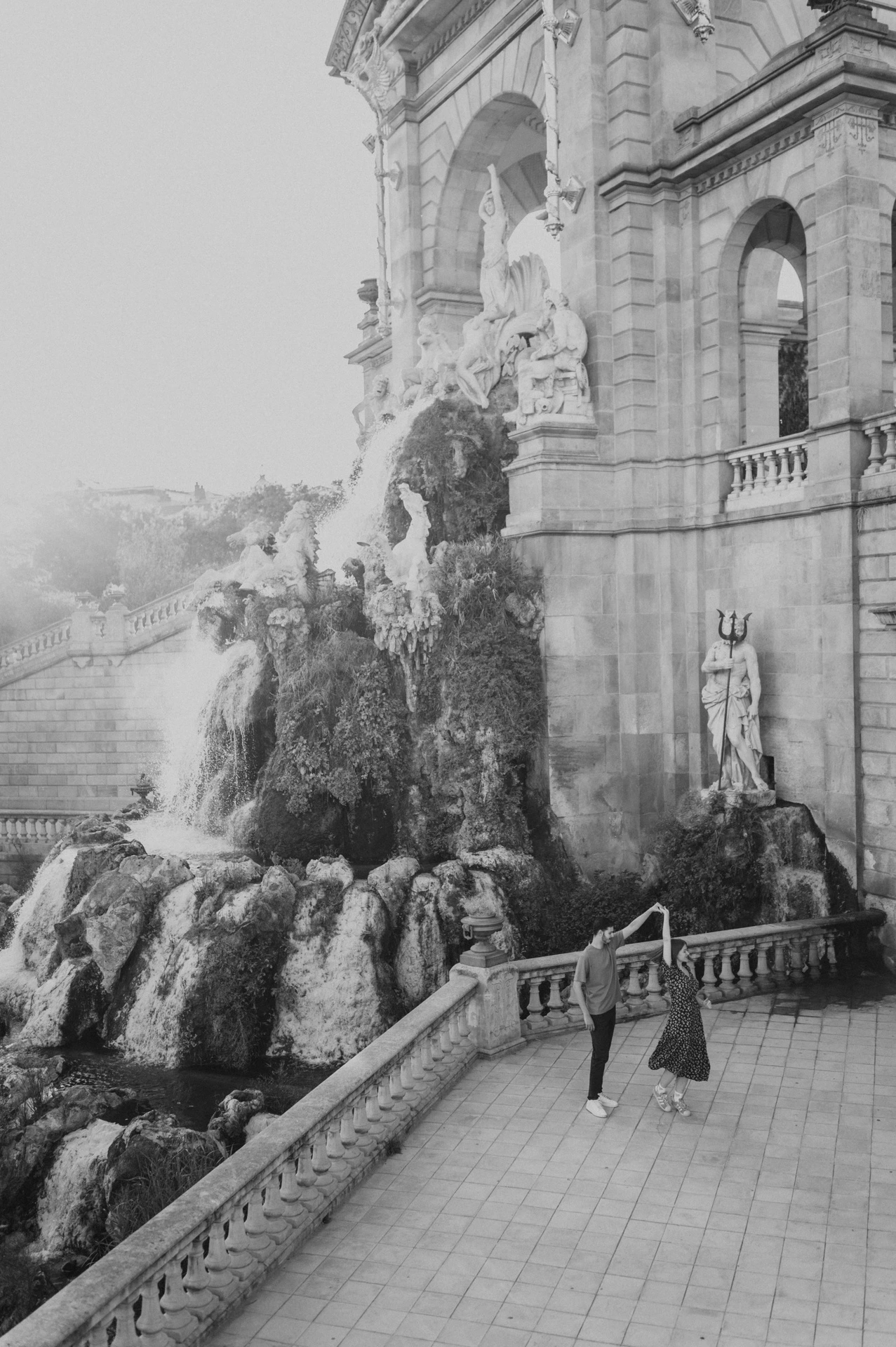 Black-and-white photograph: the couple walking hand in hand beside the fountain, with its ornate stone arches rising above them during their couple photoshoot in Barcelona.