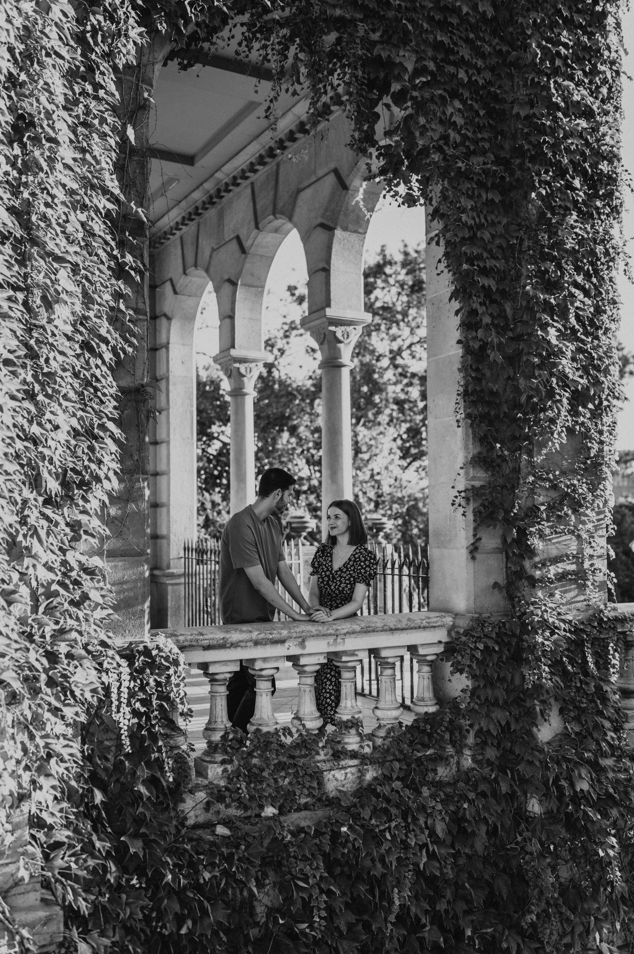 Black-and-white photograph: the couple framed by ivy and stone arches above a carved balustrade during their couple photoshoot in Barcelona.