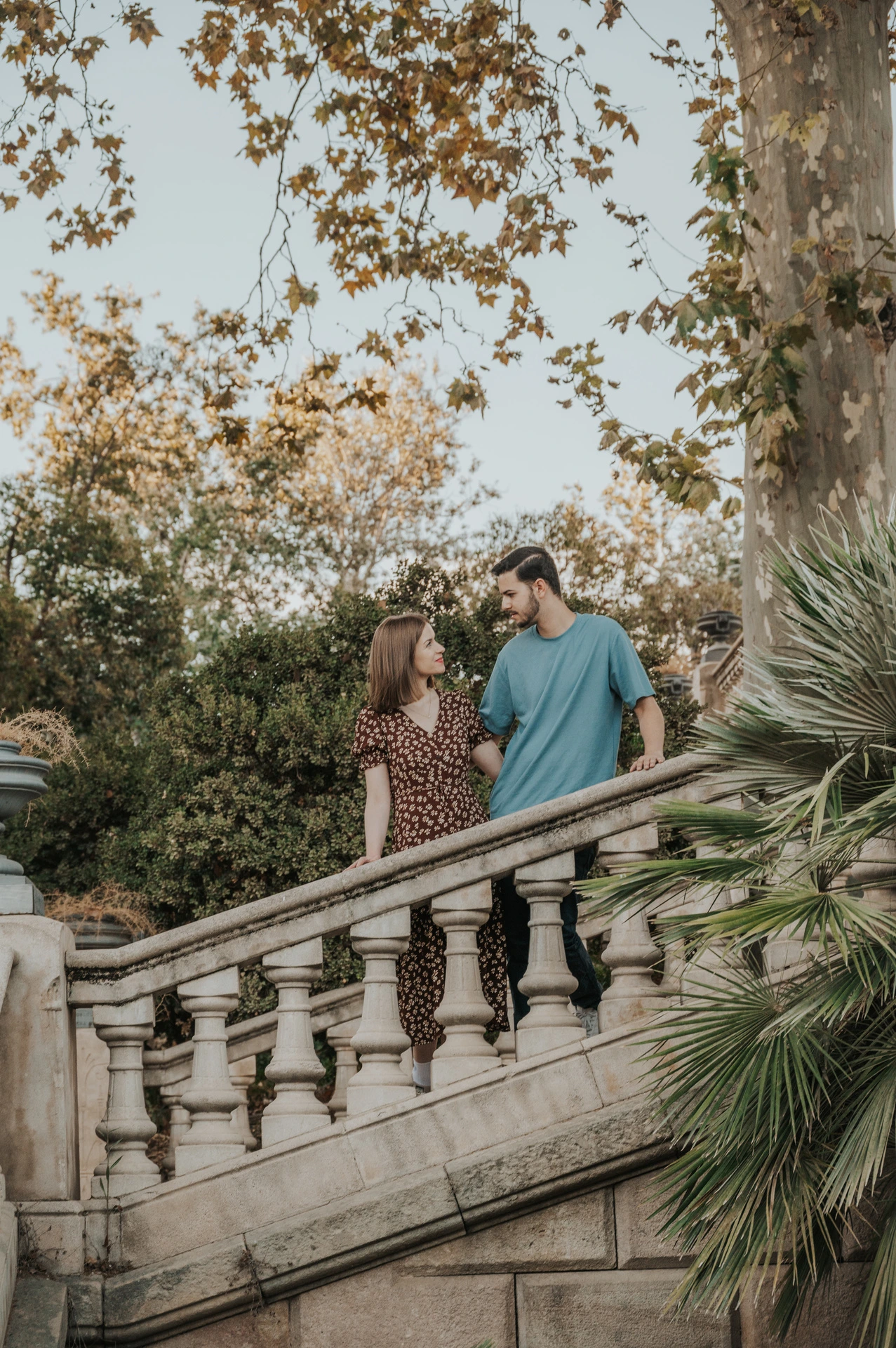 The couple standing close together on a stone staircase beneath leafy trees during their couple photoshoot in Barcelona.