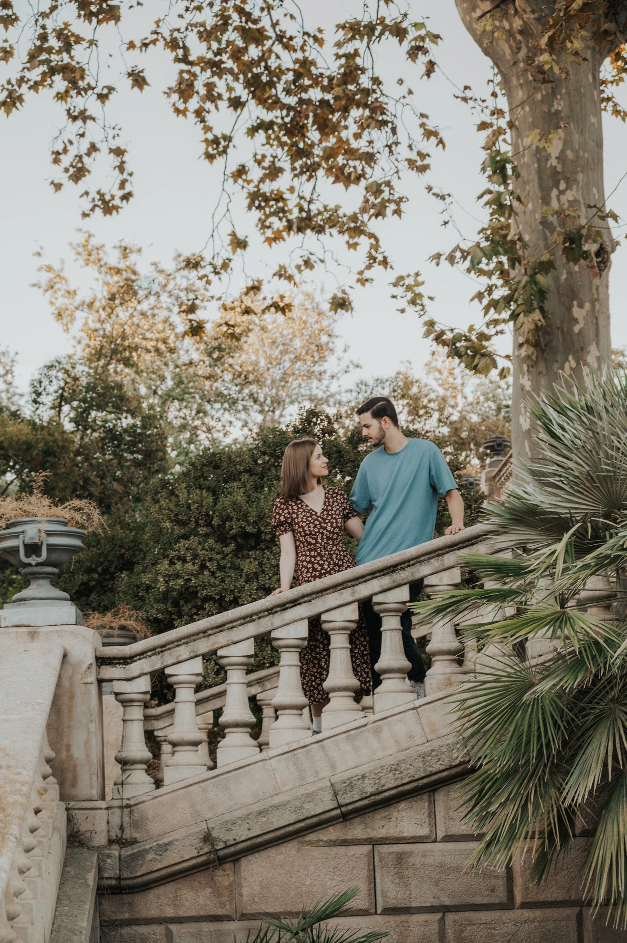 The couple standing close together on a stone staircase beneath leafy trees during their couple photoshoot in Barcelona.