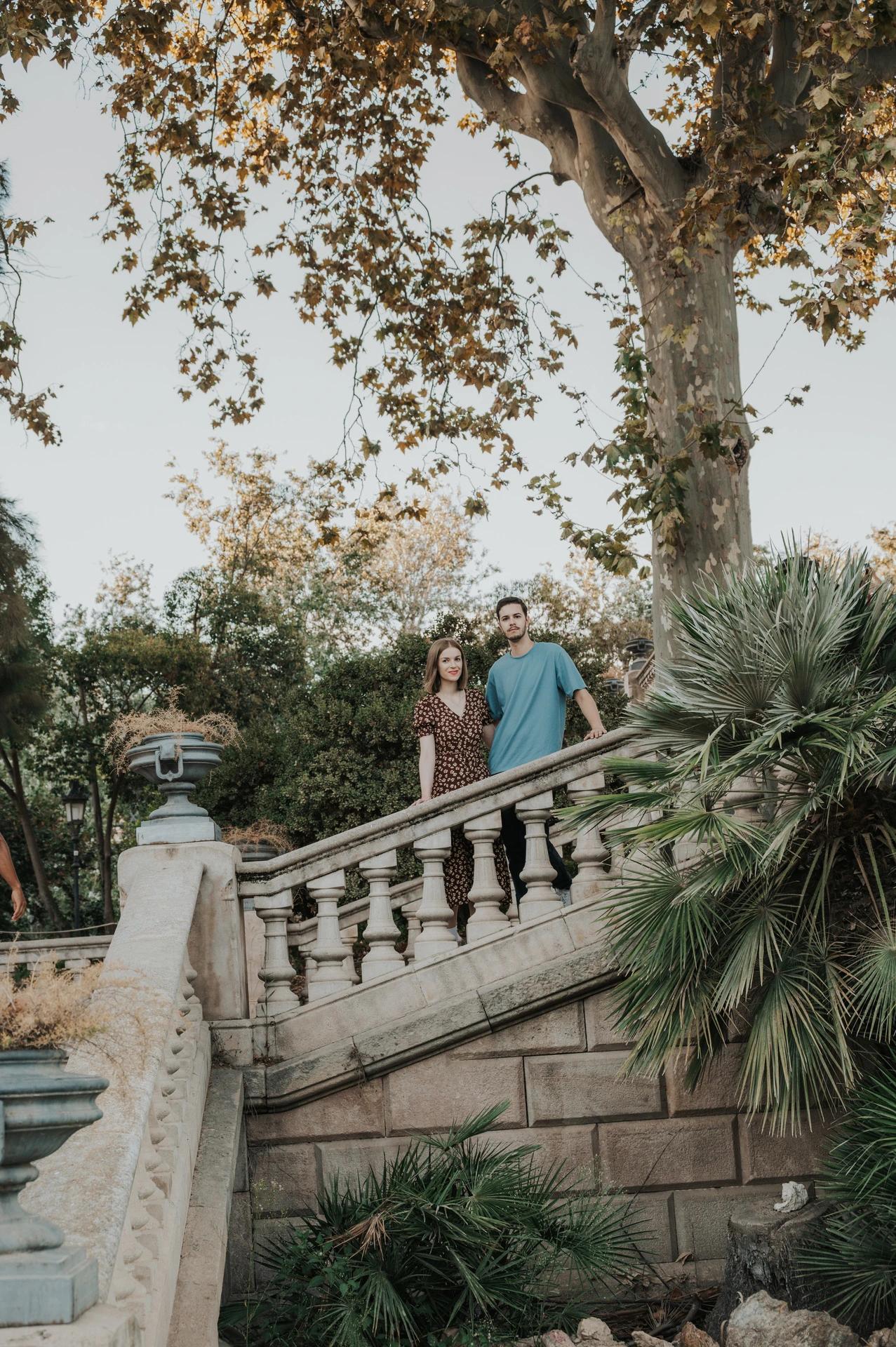 The couple standing close together on a stone staircase beneath leafy trees during their couple photoshoot in Barcelona.