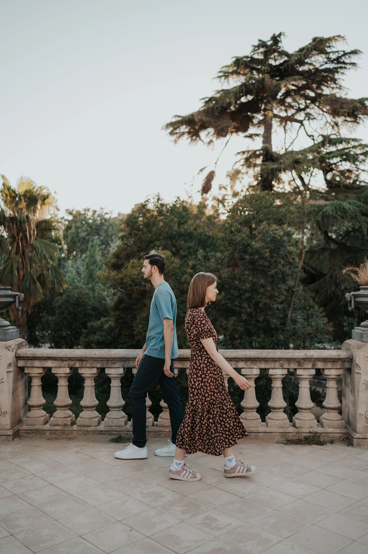 The couple walking hand in hand along a paved terrace beside trees and a stone railing during their couple photoshoot in Barcelona.