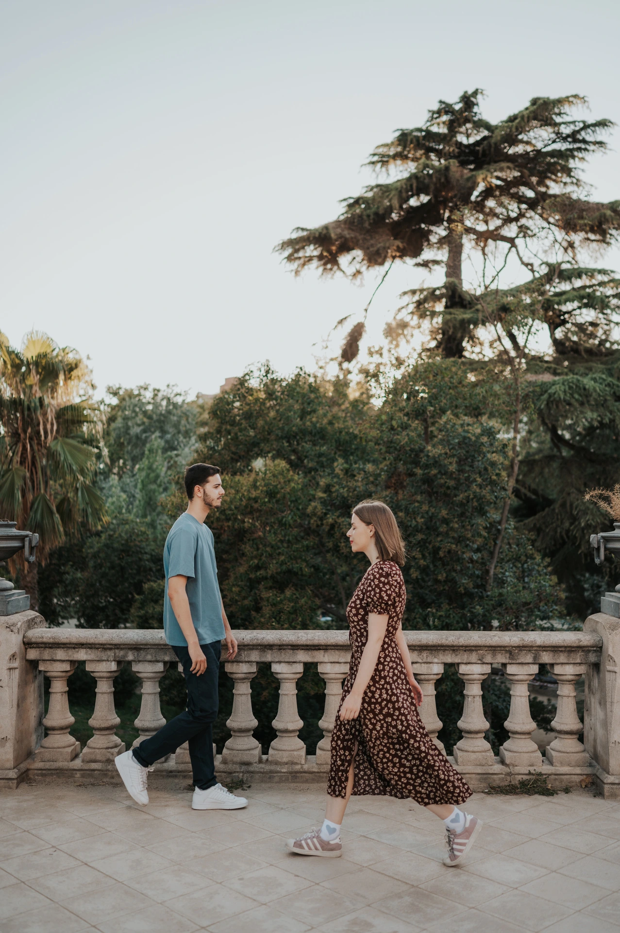 The couple walking hand in hand along a paved terrace beside trees and a stone railing during their couple photoshoot in Barcelona.