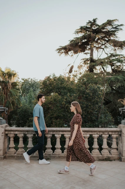 A hand-in-hand walk on the terrace | Barcelona couple photoshoot The couple walking hand in hand along a paved terrace beside trees and a stone railing during their couple photoshoot in Barcelona.